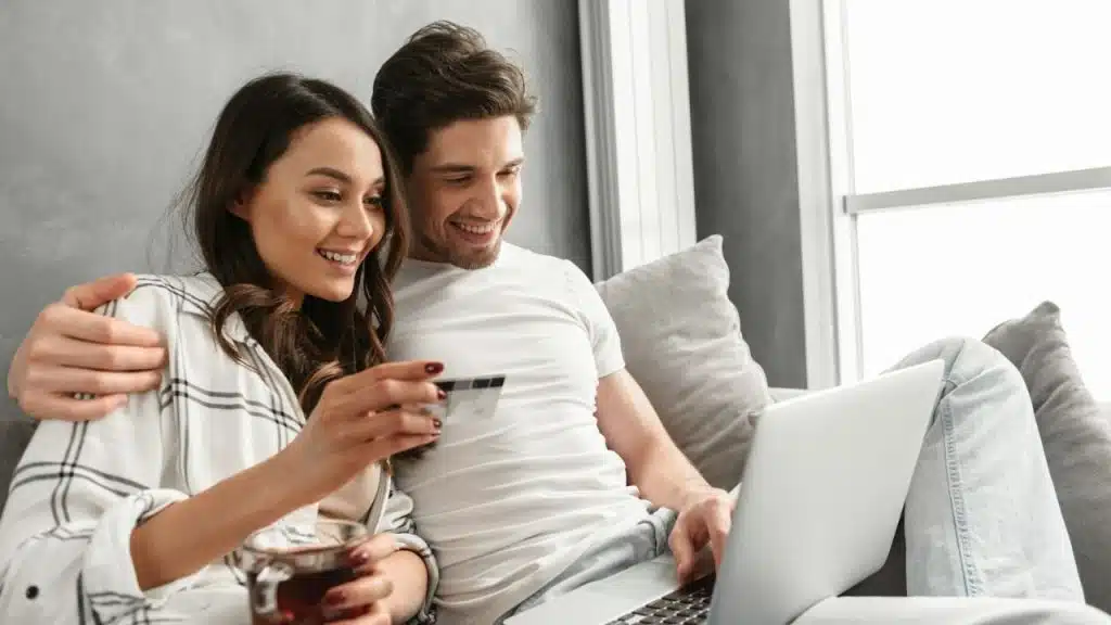 Smiling couple shopping online on a laptop, holding a credit card and tea.