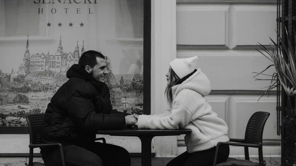 Black and white photo of a couple holding hands and smiling at a table outside a hotel.