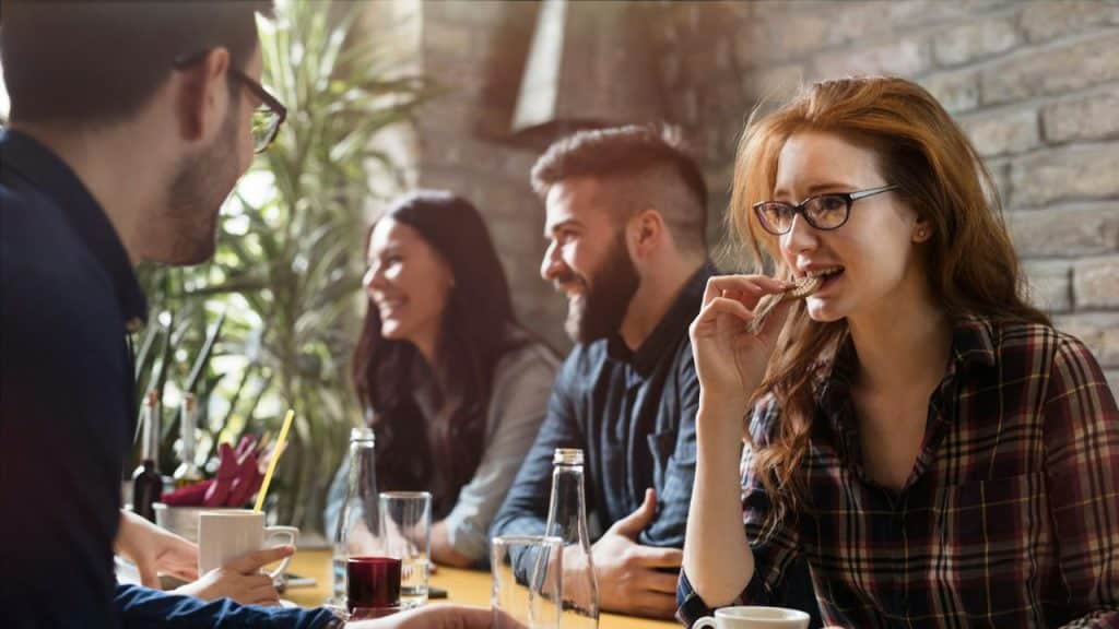 A woman in a plaid shirt and glasses bites a cookie, sitting with three smiling people.