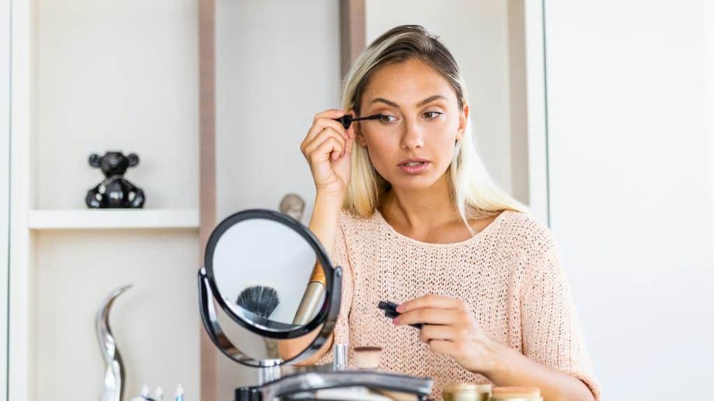 A woman applying mascara while sitting at a vanity with a mirror.
