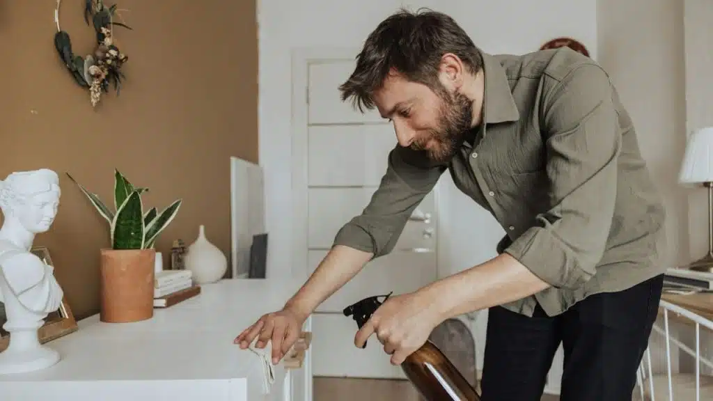 Man in an olive green shirt cleaning a white cabinet with a spray bottle and cloth.
