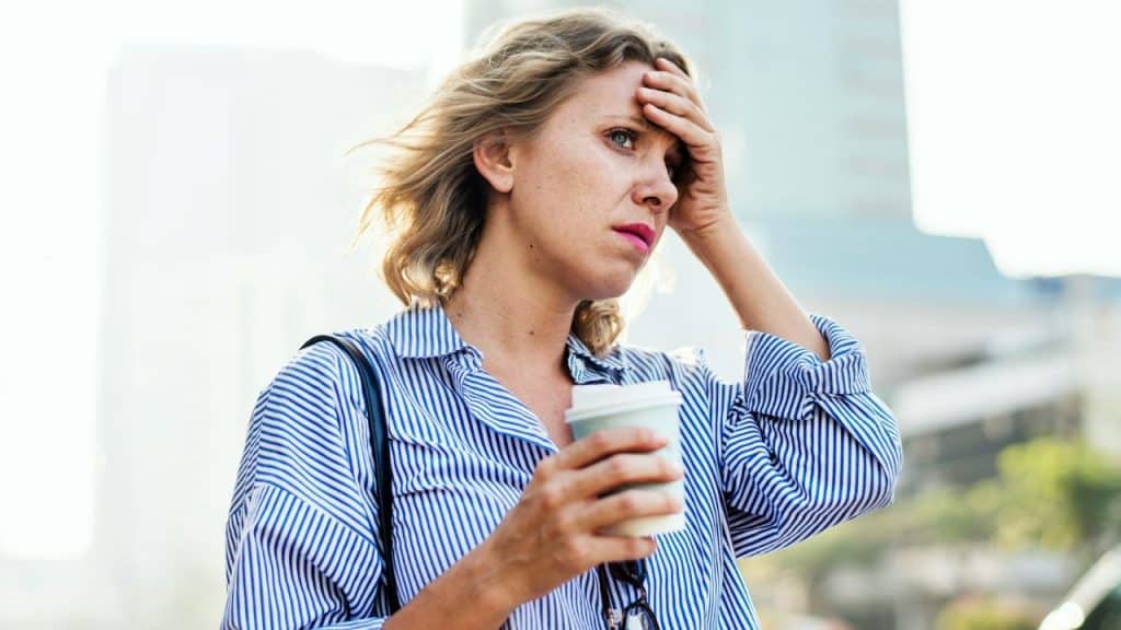 A woman holds a coffee cup and presses her hand to her forehead outdoors.
