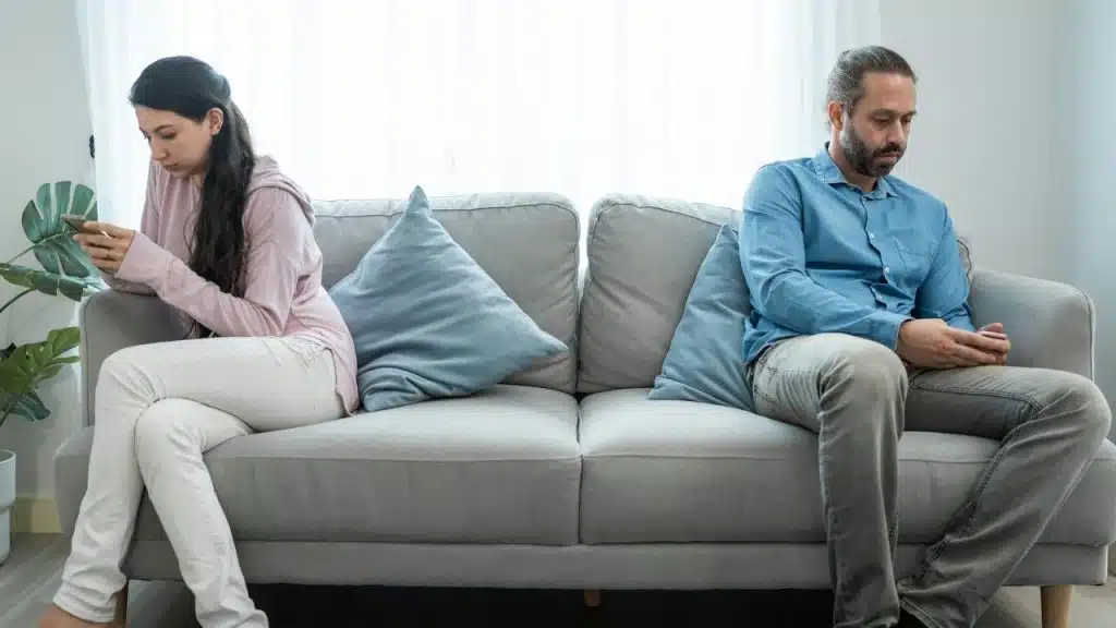 Man and woman sitting separately on a couch, facing away, both looking at phones.
