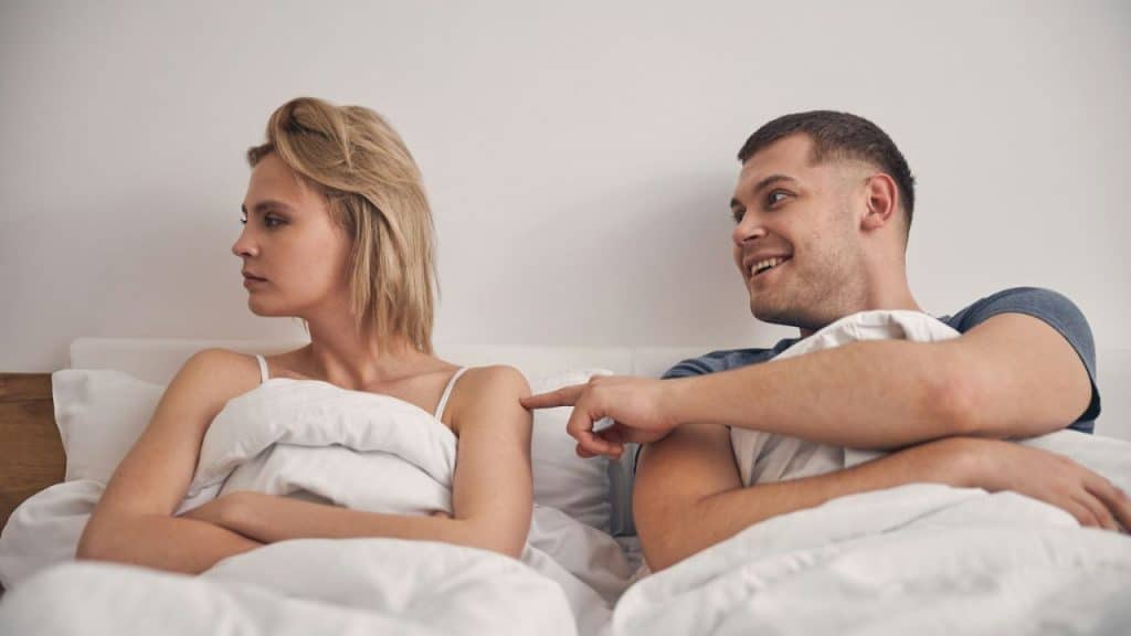 Couple in bed, woman facing away with arms crossed, man smiling and pointing.