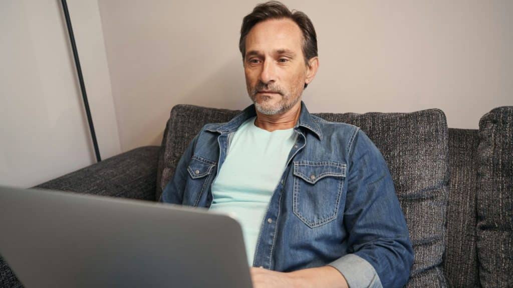A man in a denim shirt sits on a sofa, looking intently at a laptop screen.