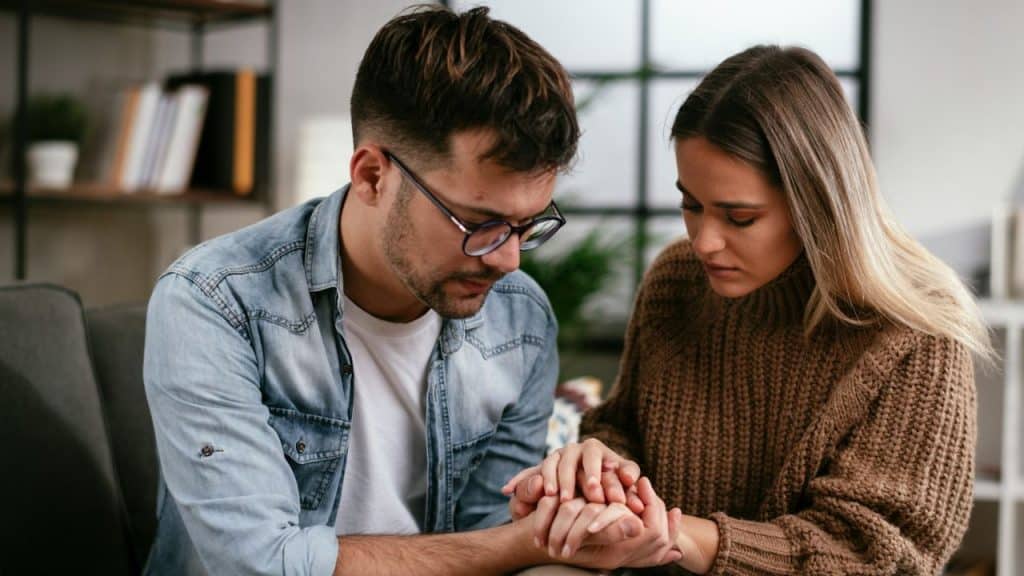 Man in glasses and woman holding hands, looking down seriously in a living room.