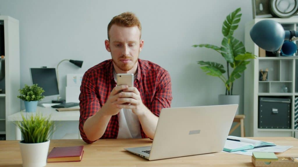 A red-haired man in a plaid shirt sits at a desk, looking down at his smartphone.