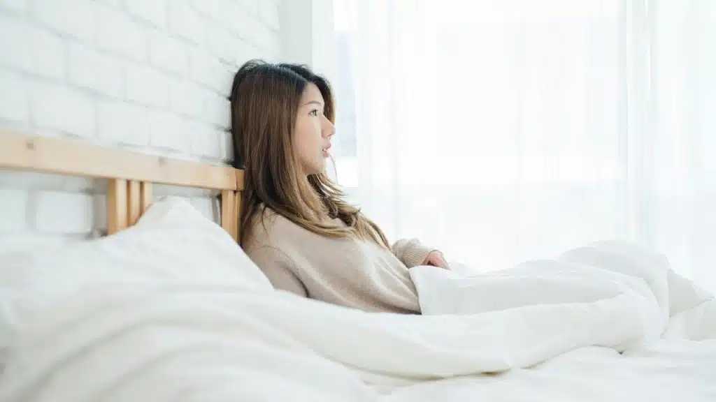 A woman sitting up in bed, leaning against a white brick wall and looking out the window.