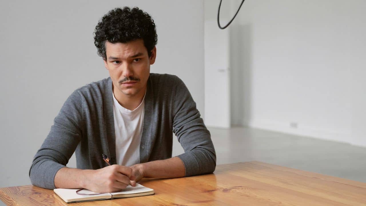 A man with curly hair and a mustache sits at a table, writing in a notebook.