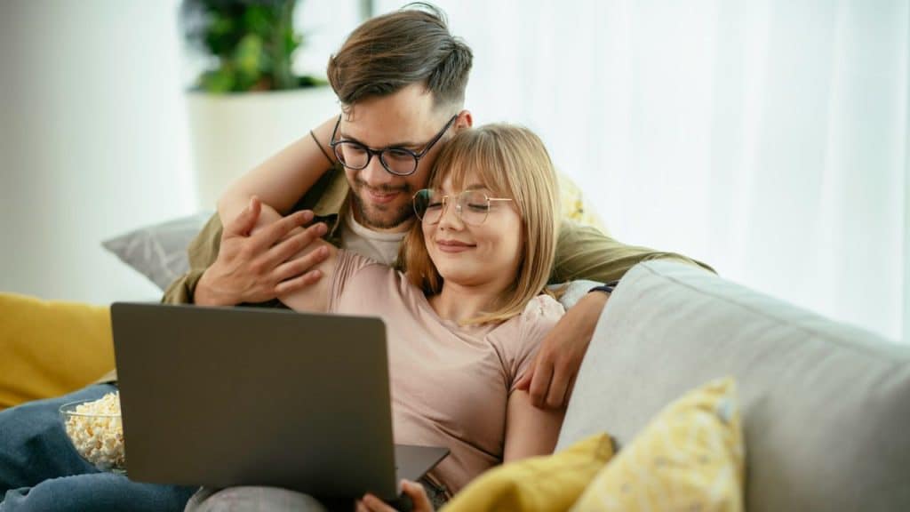 Smiling man and woman on a leather couch, looking at a laptop together indoors.