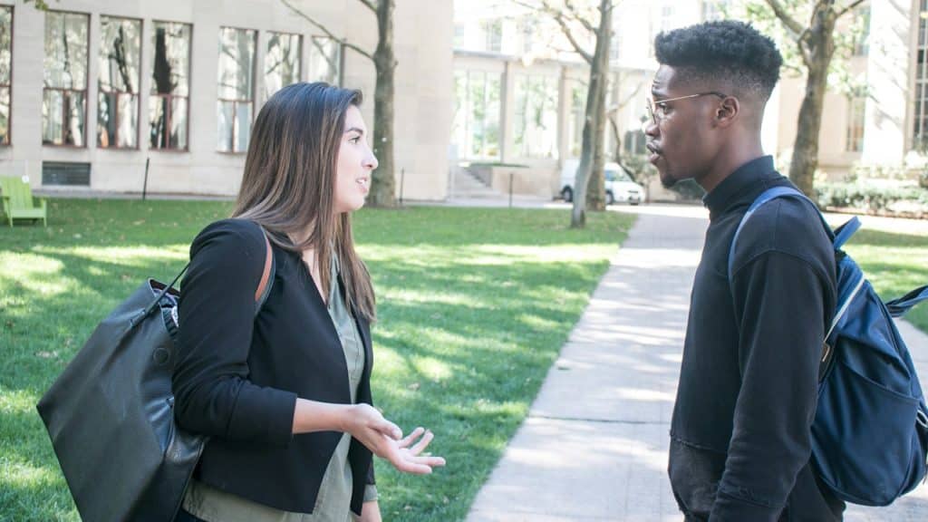 Two students, a woman and a man, talking outdoors on a university campus path.