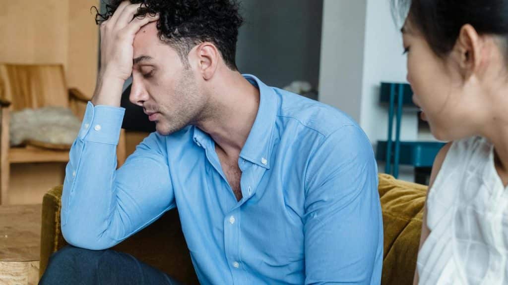 A man holding his head while sitting beside another person indoors.