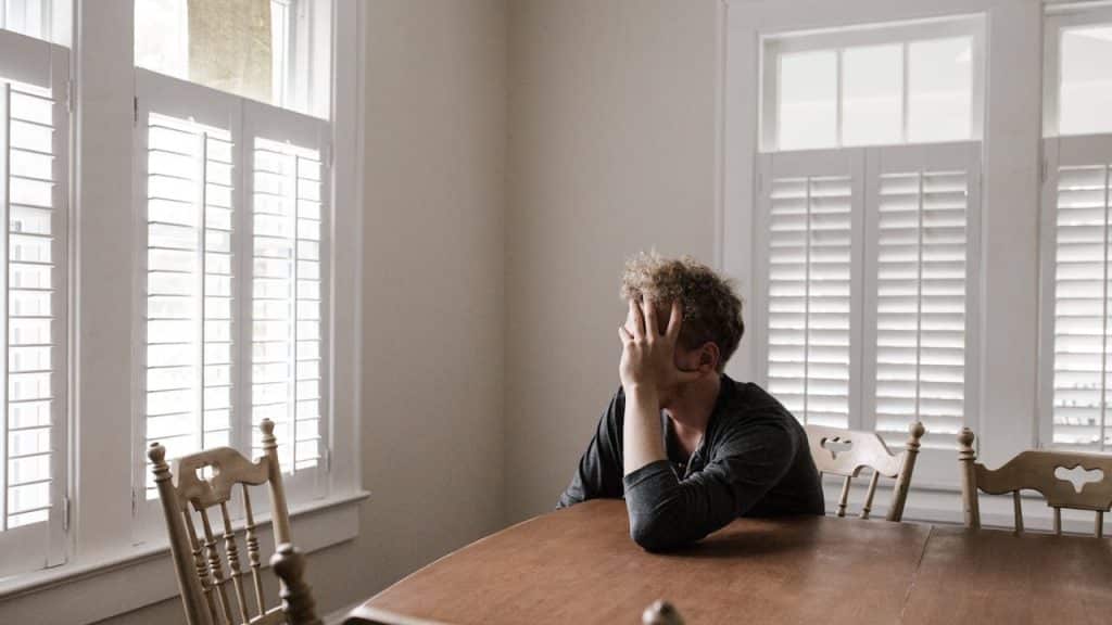 A man sits at a wooden table in a room with large windows and shutters, covering his face with his hand in distress.