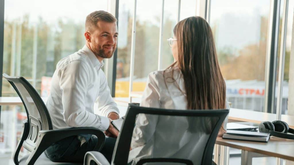 Man and woman in office chairs talking face-to-face near a sunny window.