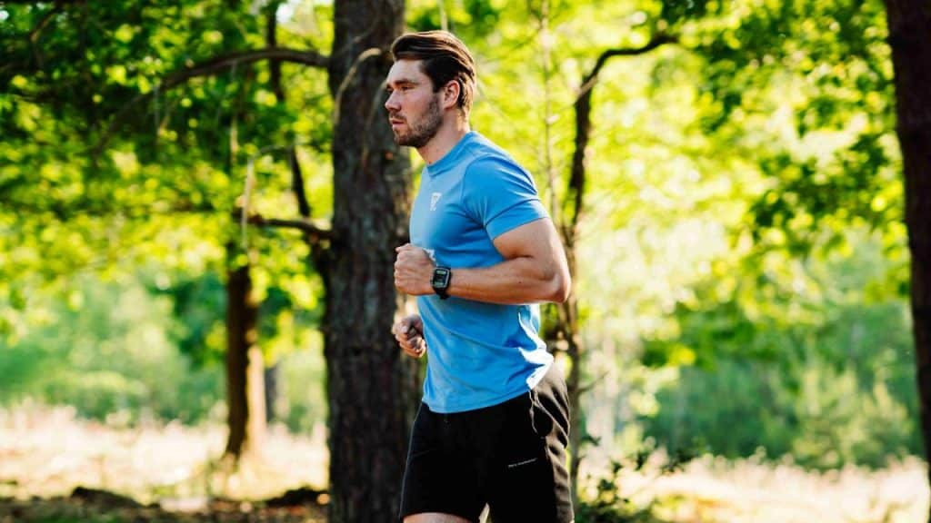 Man in blue shirt and black shorts running on a trail in a sunlit green forest.