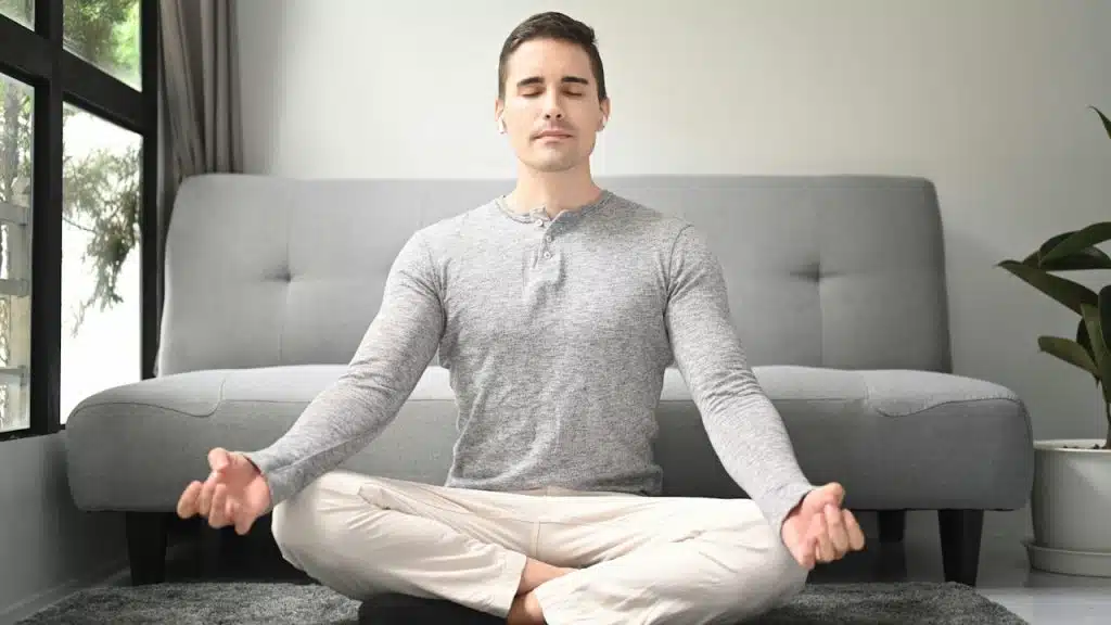 Man in lotus position meditating on a rug in a living room.