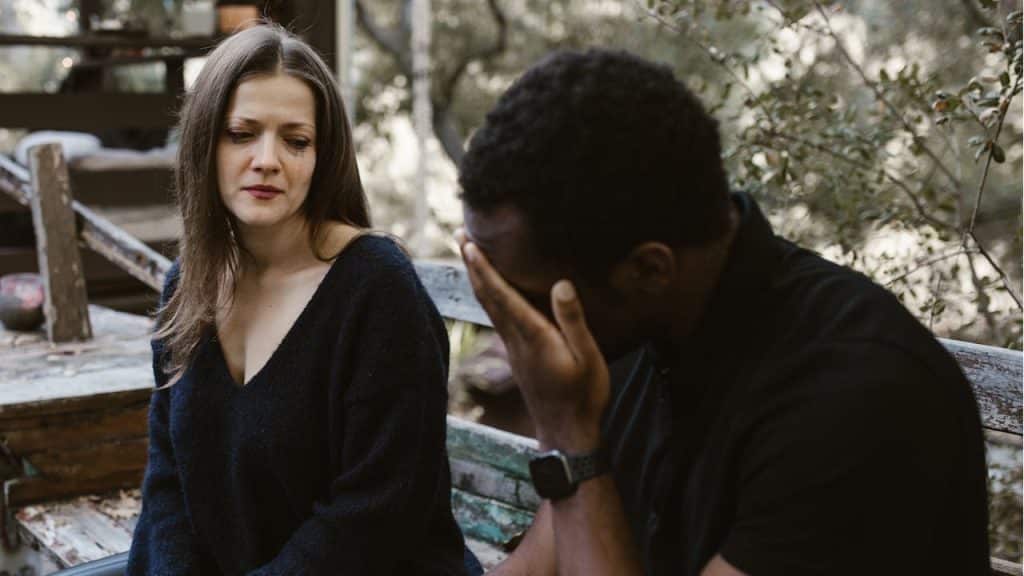 A woman looks concerned as she sits next to a distressed man holding his head.