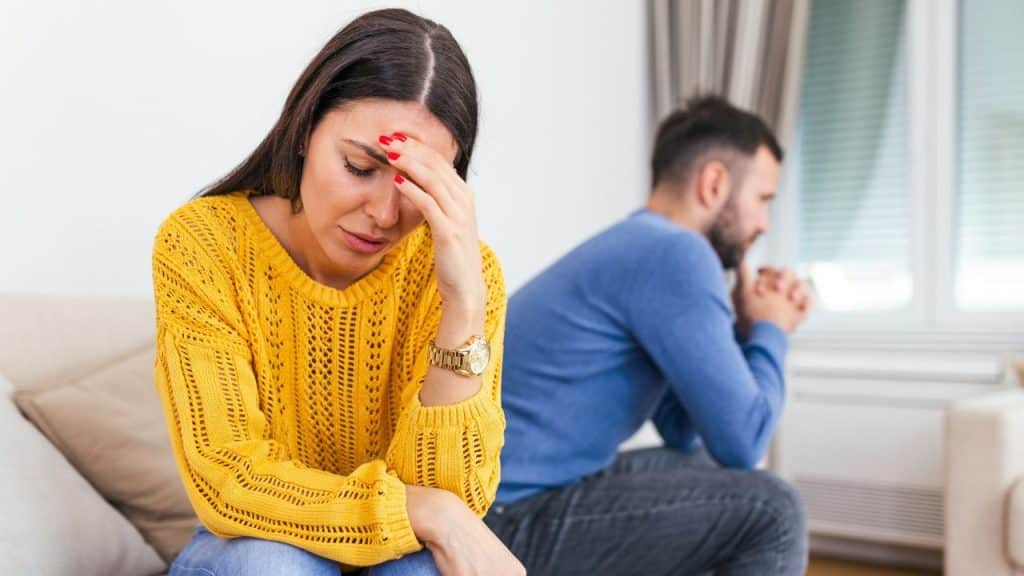 A woman in a yellow sweater holds her head, while a man sits nearby in the background, looking away.