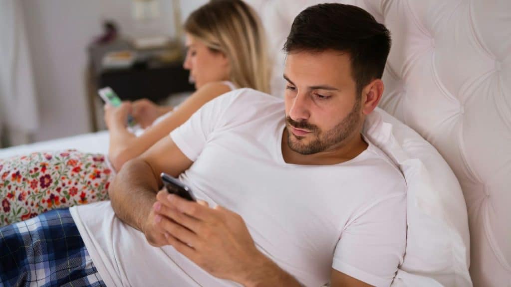 Man in white shirt texting on phone in bed; woman in background uses her phone.