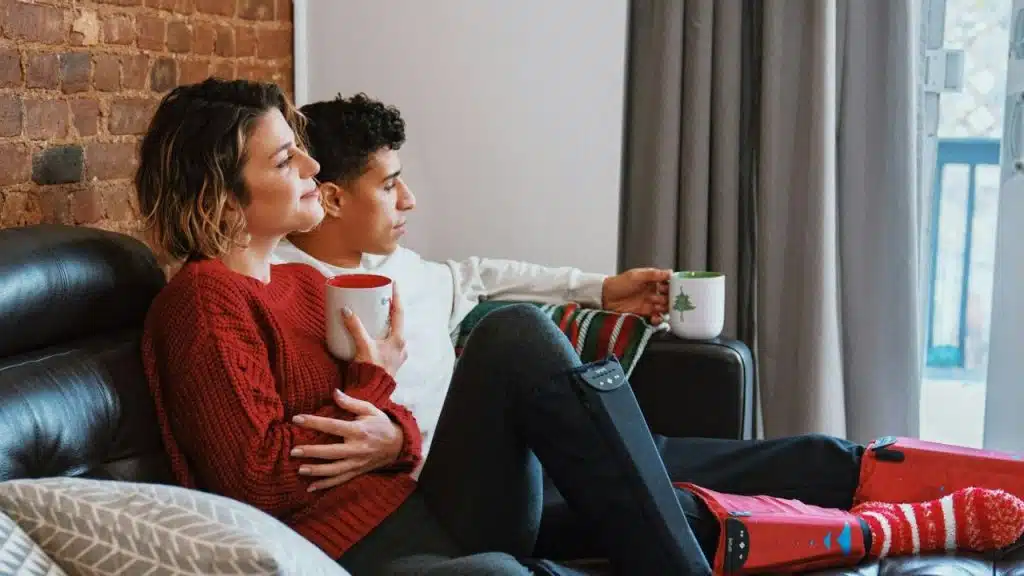 A couple sits on a dark couch holding mugs, looking toward a large window on the right.