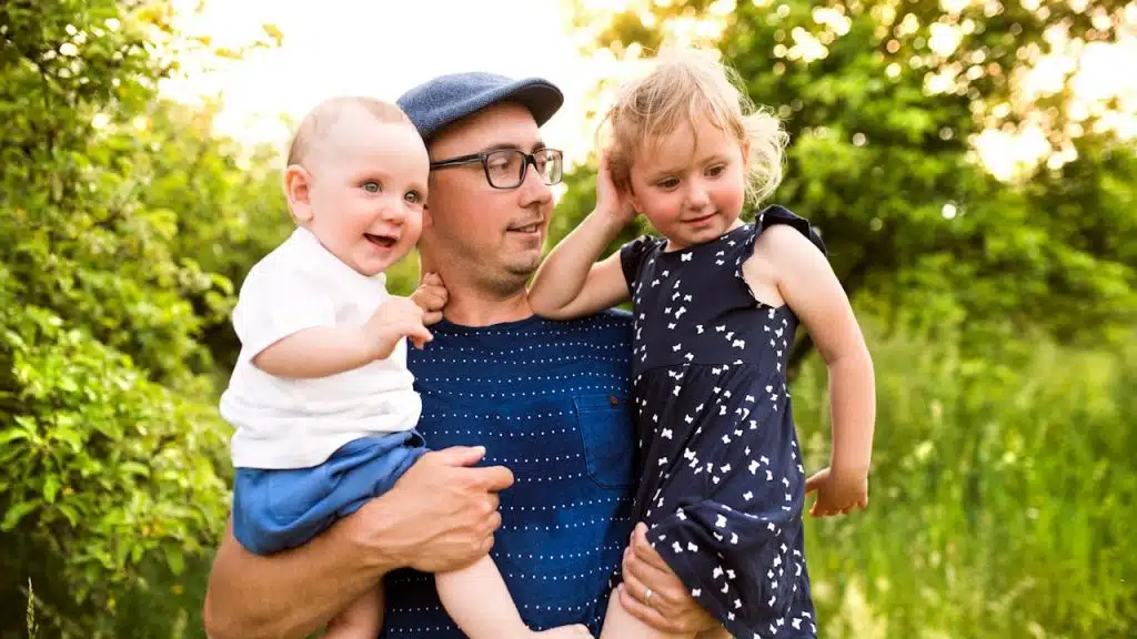 A father in a blue cap and glasses holds a smiling baby and a small girl in a dark dress outdoors in a sunny, green setting.