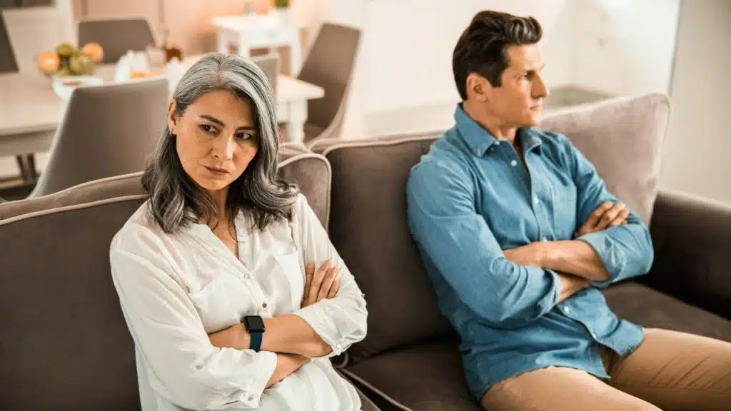 A middle-aged woman and man sit separated on a couch, both with arms crossed.