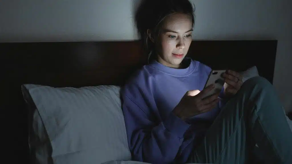 A woman sitting in bed at night looking at her phone.
