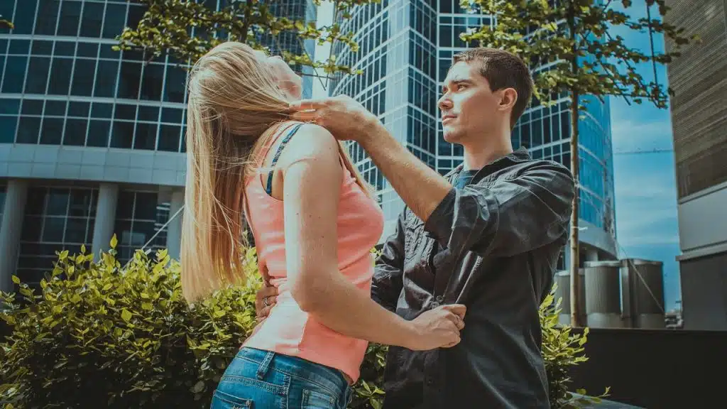 A man in a dark shirt gently touches a woman's neck outdoors near skyscrapers and green foliage.