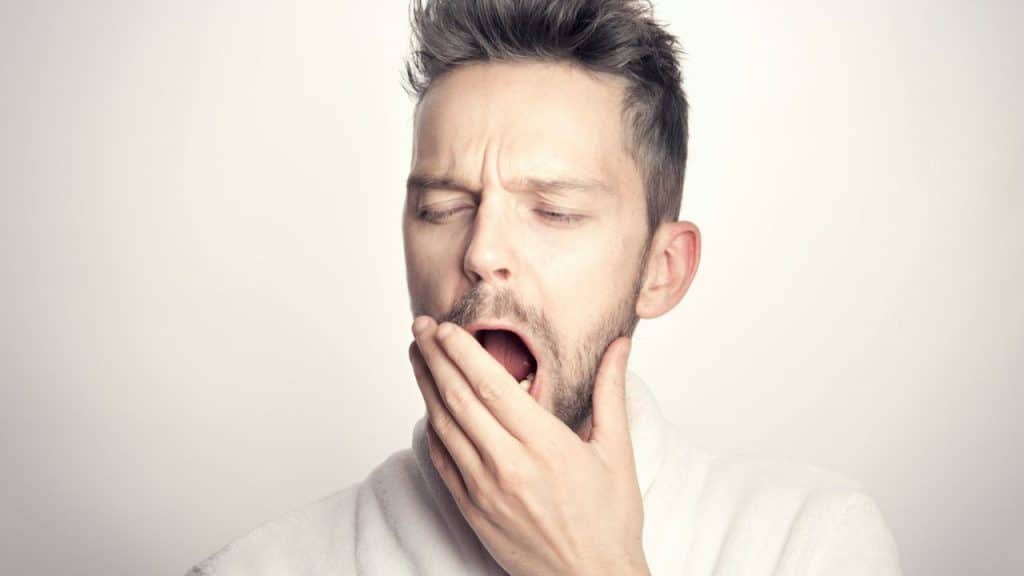 A man in a white robe yawns widely, covering his mouth with his hand.