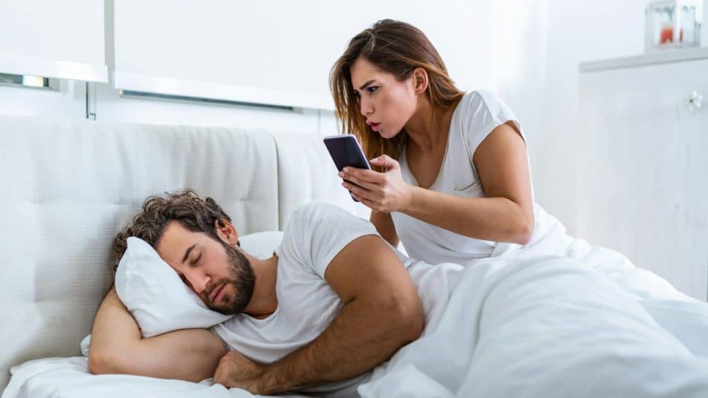 A woman in a white t-shirt sits on a bed looking at a phone while a man with a beard sleeps next to her.