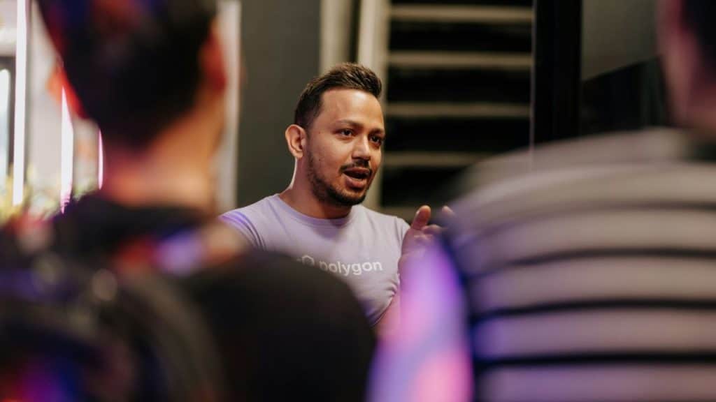 Man in a light purple Polygon t-shirt talking to people in a dark, indoor setting.
