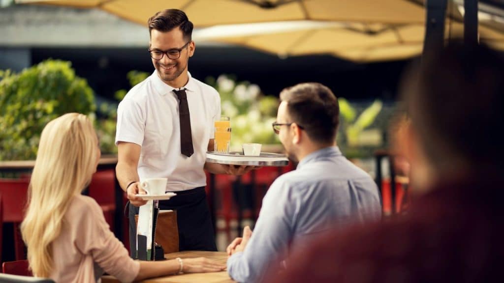 Smiling waiter in a white shirt serving coffee and juice to a couple at a table.
