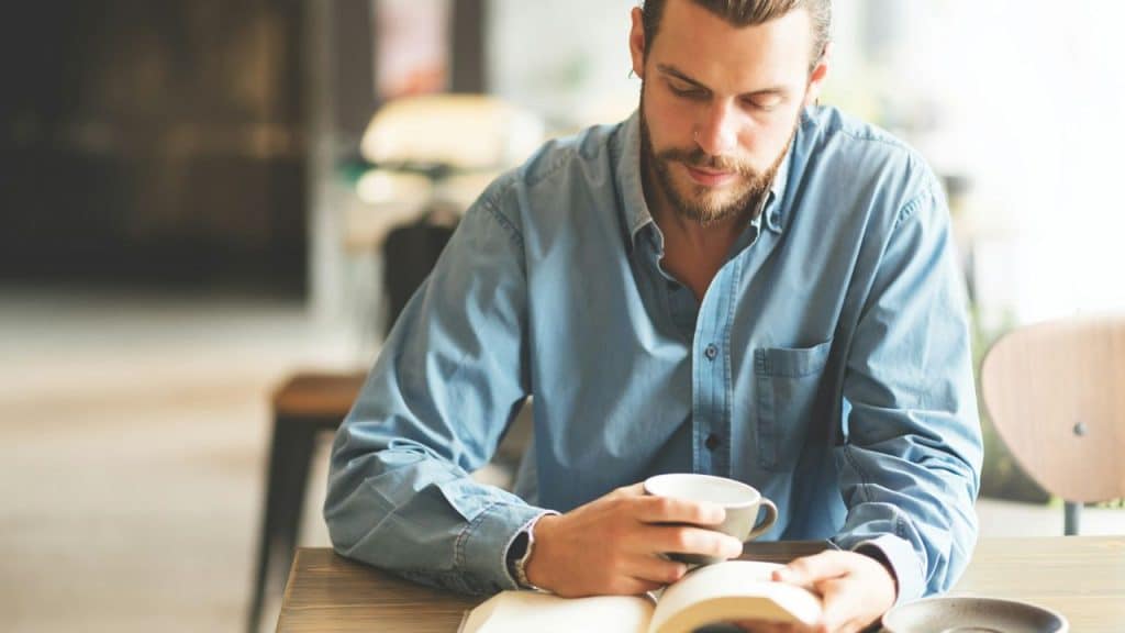 A man with a beard sits alone at a wooden table reading a book and holding a mug.