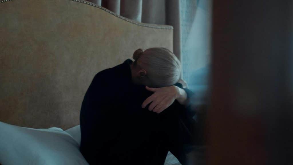 A person sitting on a bed with head bowed and arm covering their face in a dimly lit room.