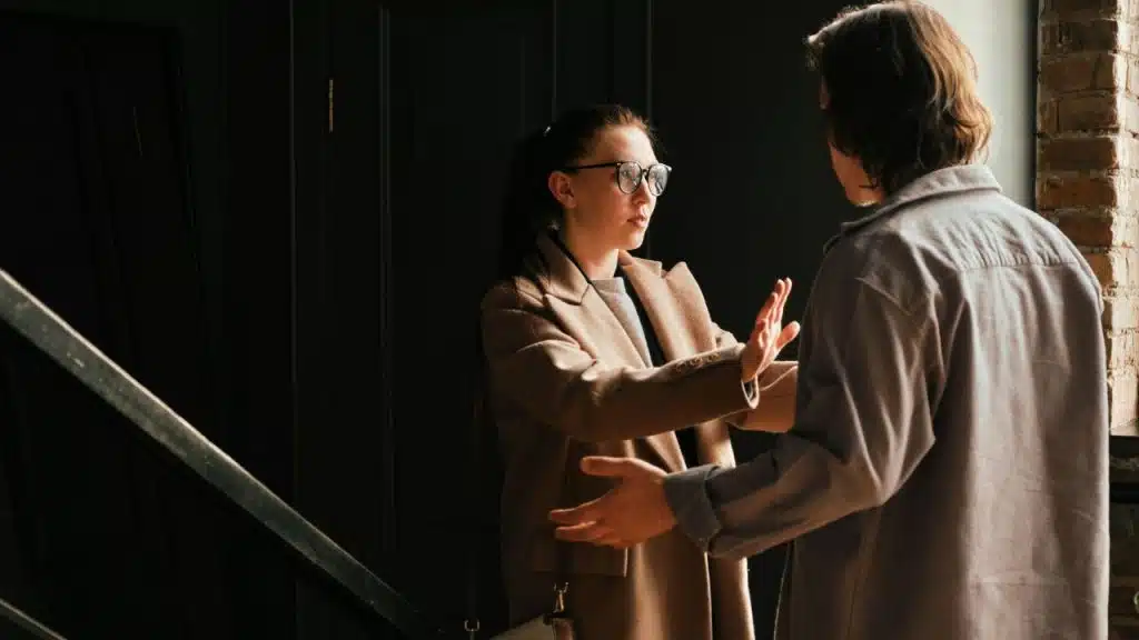 A woman in a coat holds her hand up toward a man standing in a doorway.