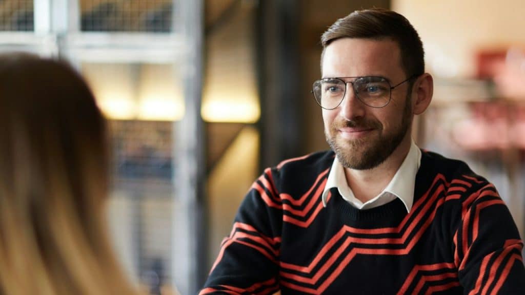 A man with glasses and a beard smiles while looking at a woman in profile.