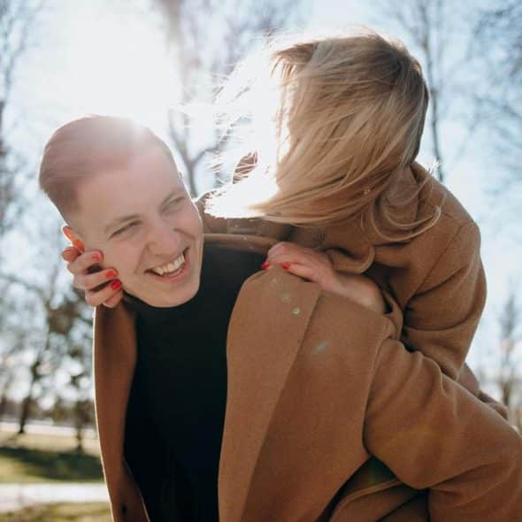 A smiling person giving another a piggyback ride outdoors in sunlight.