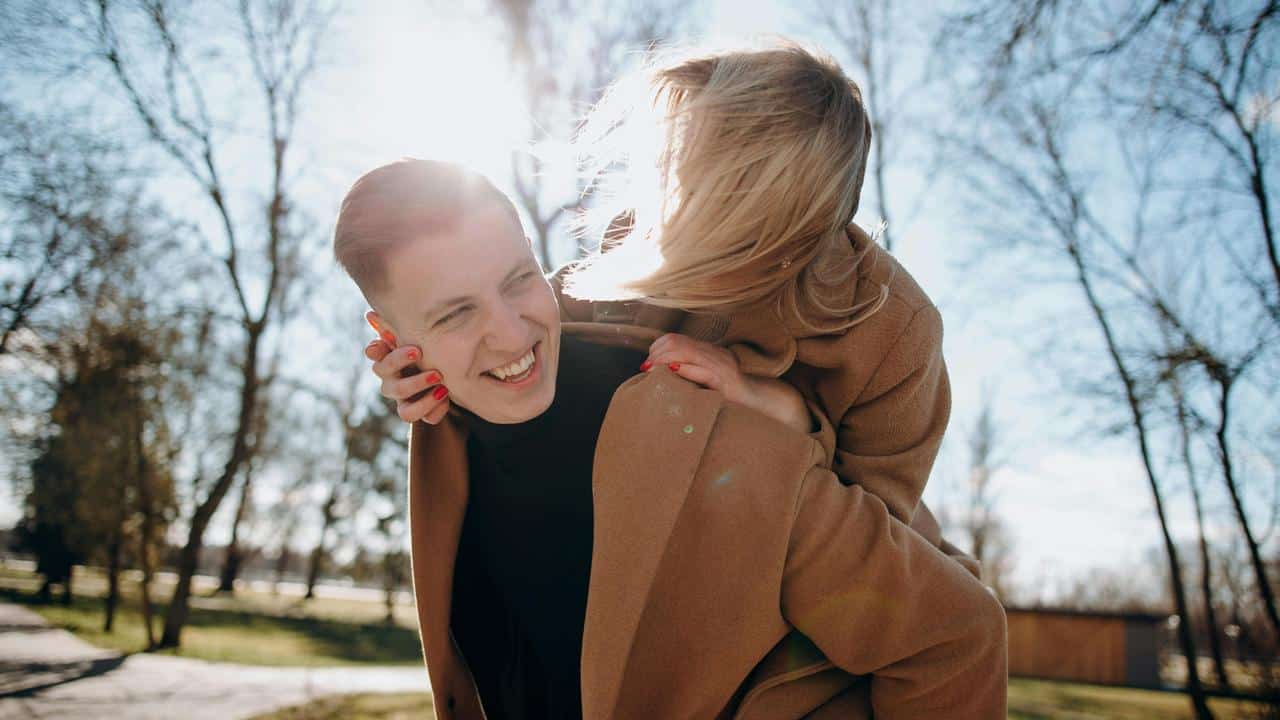 A smiling person giving another a piggyback ride outdoors in sunlight.