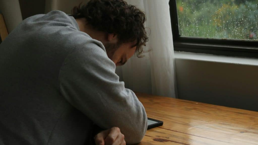 A man with curly hair sits at a wooden table looking down near a rainy window.