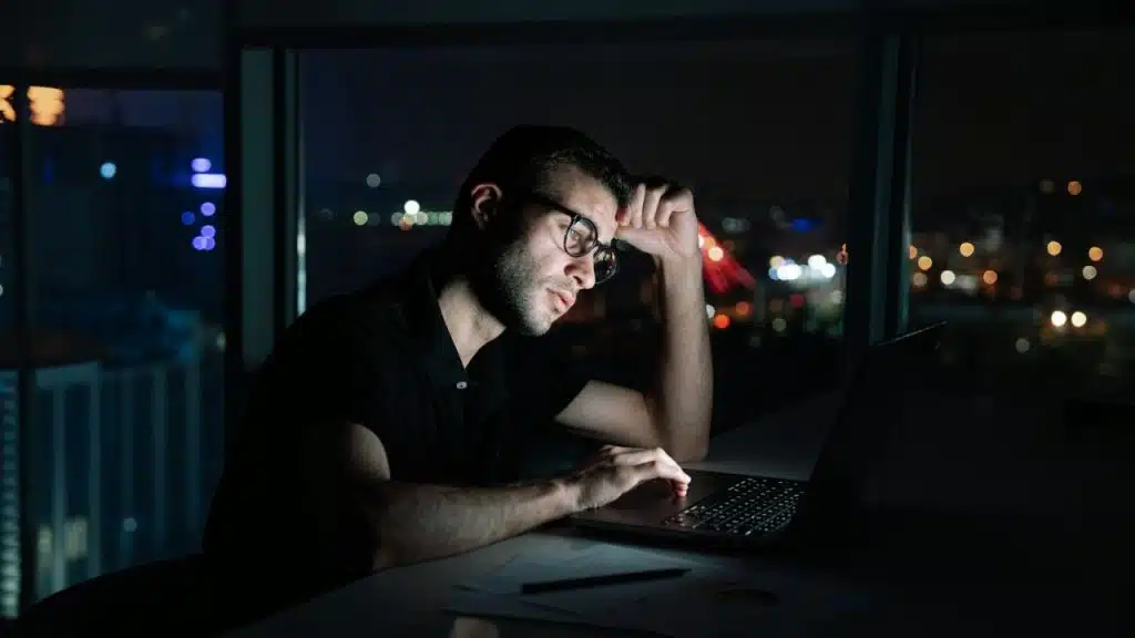 A man in a black polo shirt and glasses works on a laptop at night.