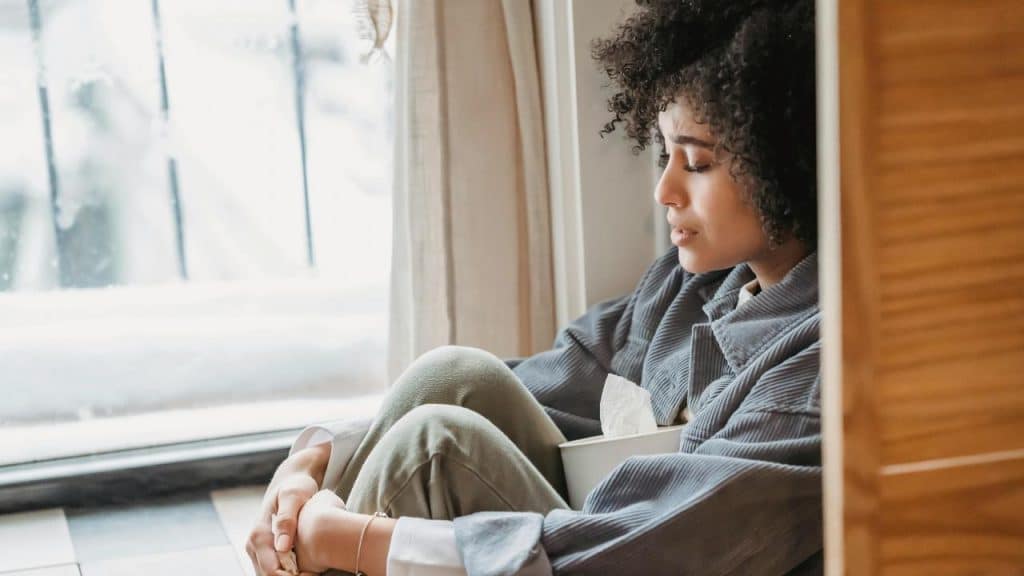 A woman sitting by a window with her knees drawn up, holding a tissue box and looking down.