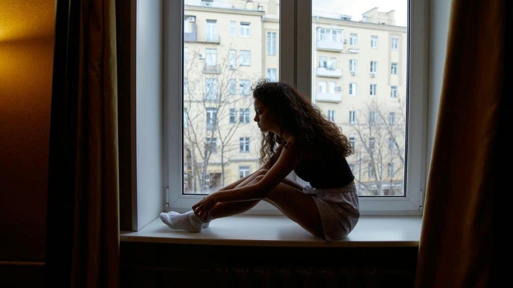 A woman sitting on a wide windowsill, looking outside thoughtfully.