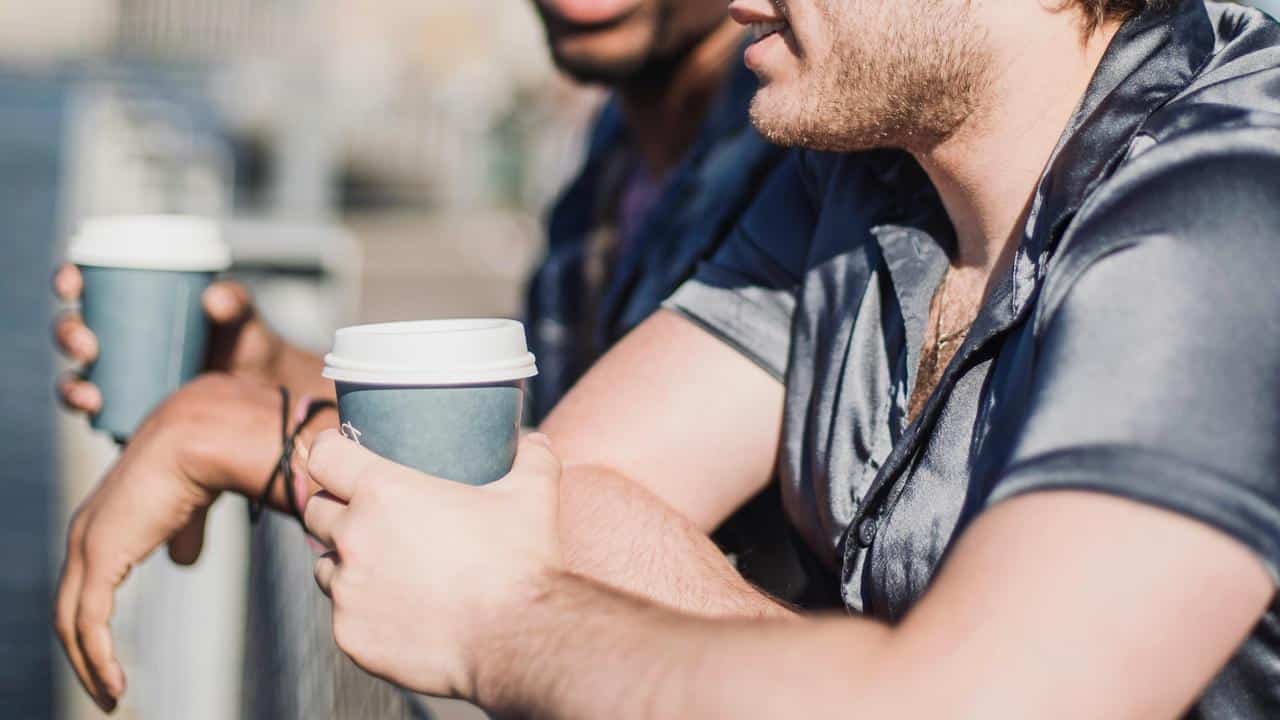 A close-up of two men holding takeaway coffee cups while talking outdoors.