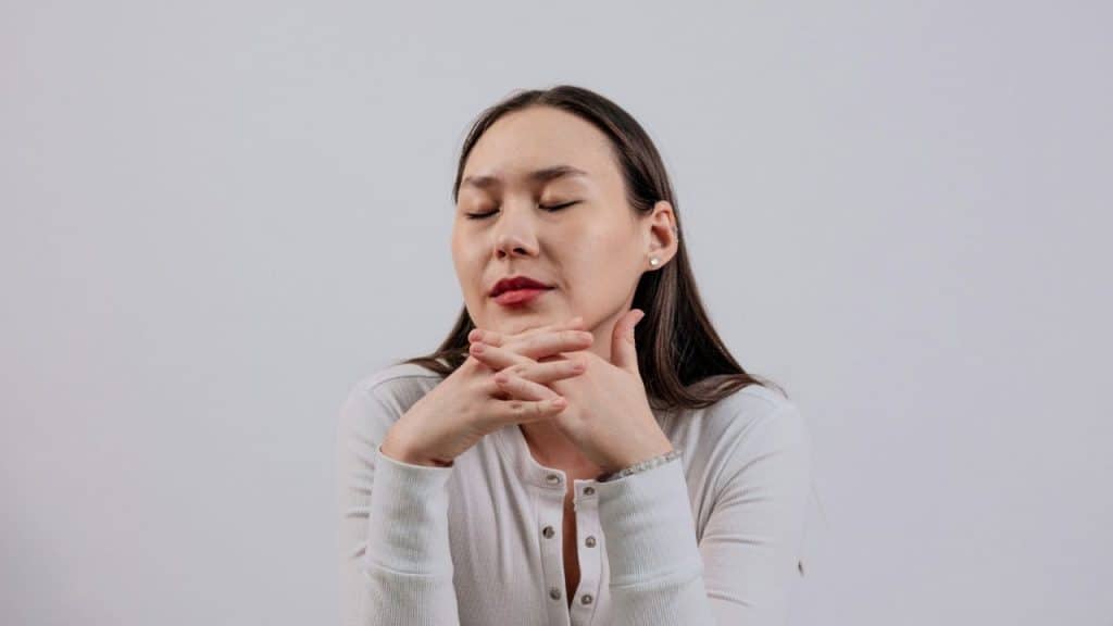 A woman with closed eyes rests her chin on her interlaced fingers against a plain background.