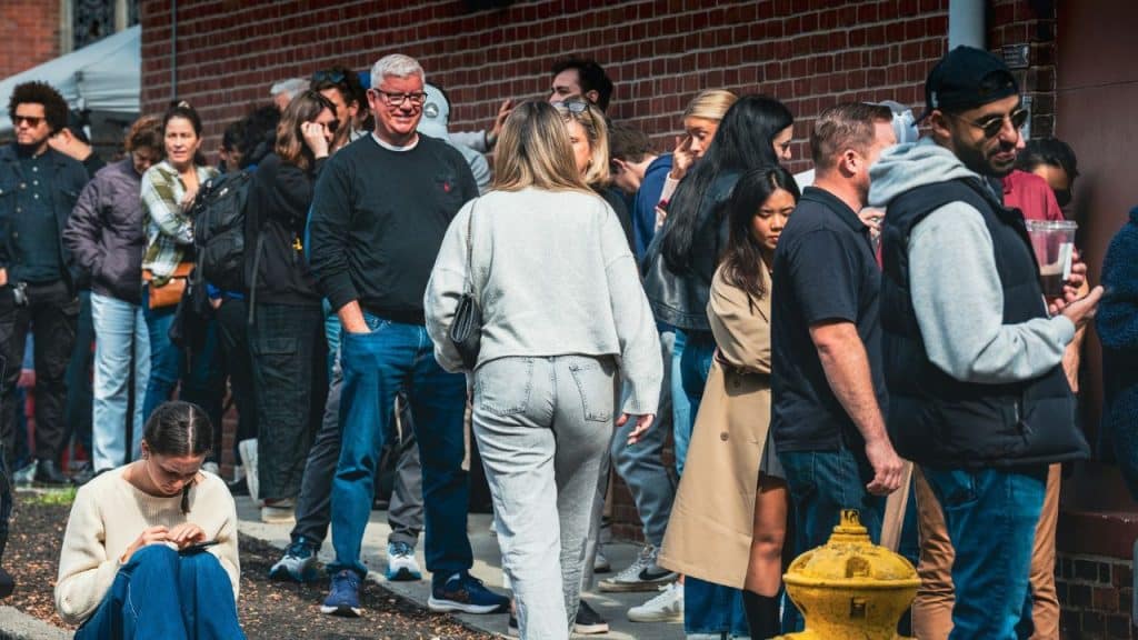 A diverse group of people stands in a long line along a brick building.
