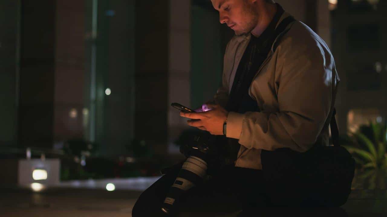 A man sitting outdoors at night checking his phone with a camera resting on his lap.