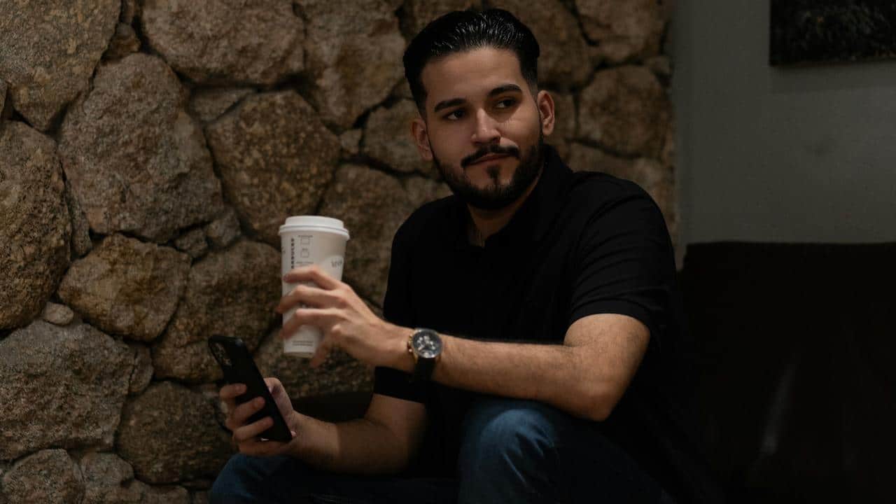 A man sitting indoors holding a coffee cup and a phone.