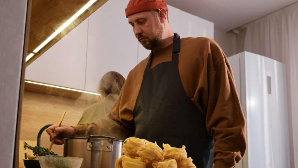 Man in a beanie and apron cooking pasta in a modern kitchen.