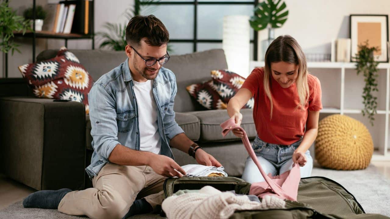 Smiling couple sitting on the floor together and packing a large suitcase in a living room.