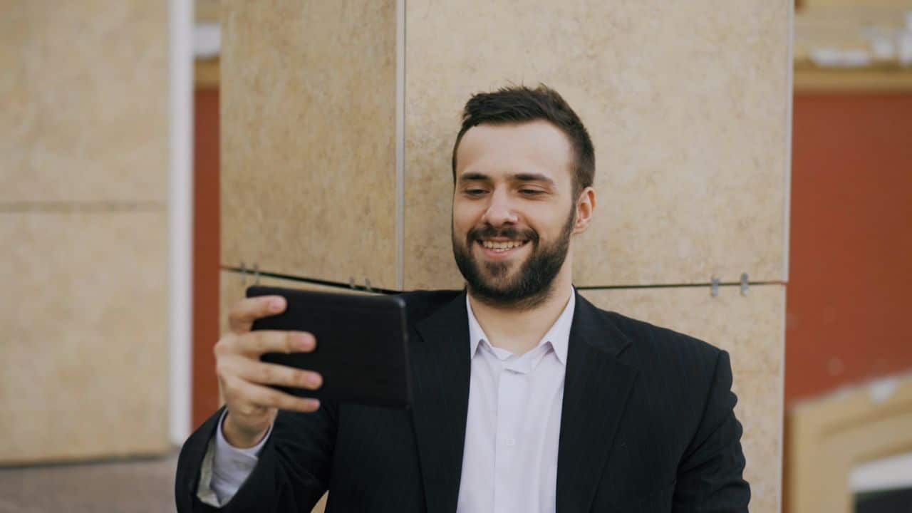 Smiling bearded man in a suit jacket holding and looking at a black tablet outdoors.